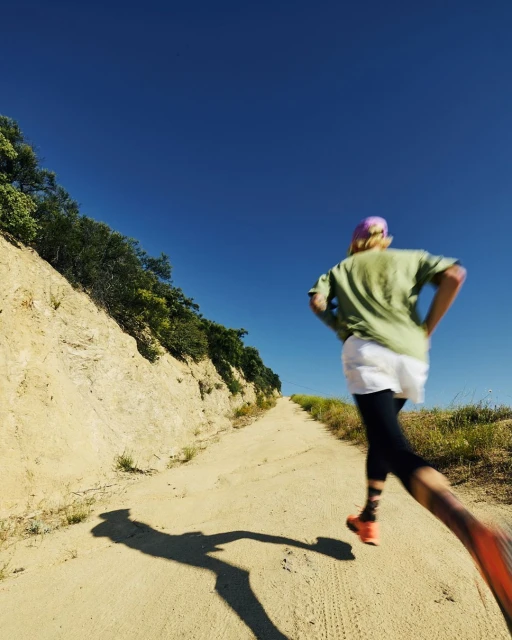 Woman trail running outdoors on a dirt path under a clear blue sky, captured in a sunny, wide shot, focused on fitness and nature.