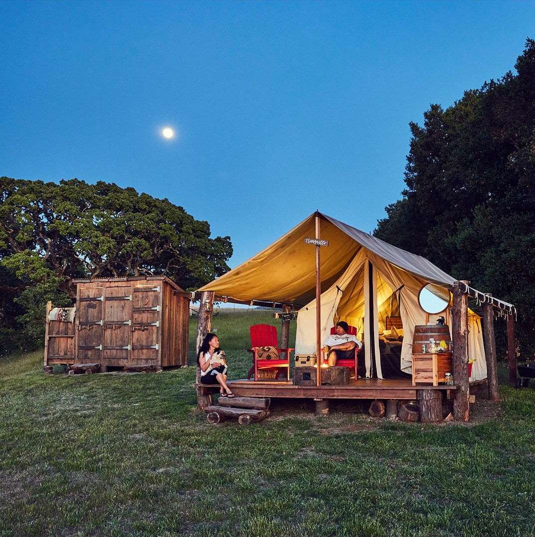 Two people relax outside a canvas tent with wooden platform and steps at dusk under a night sky with a full moon, surrounded by trees.