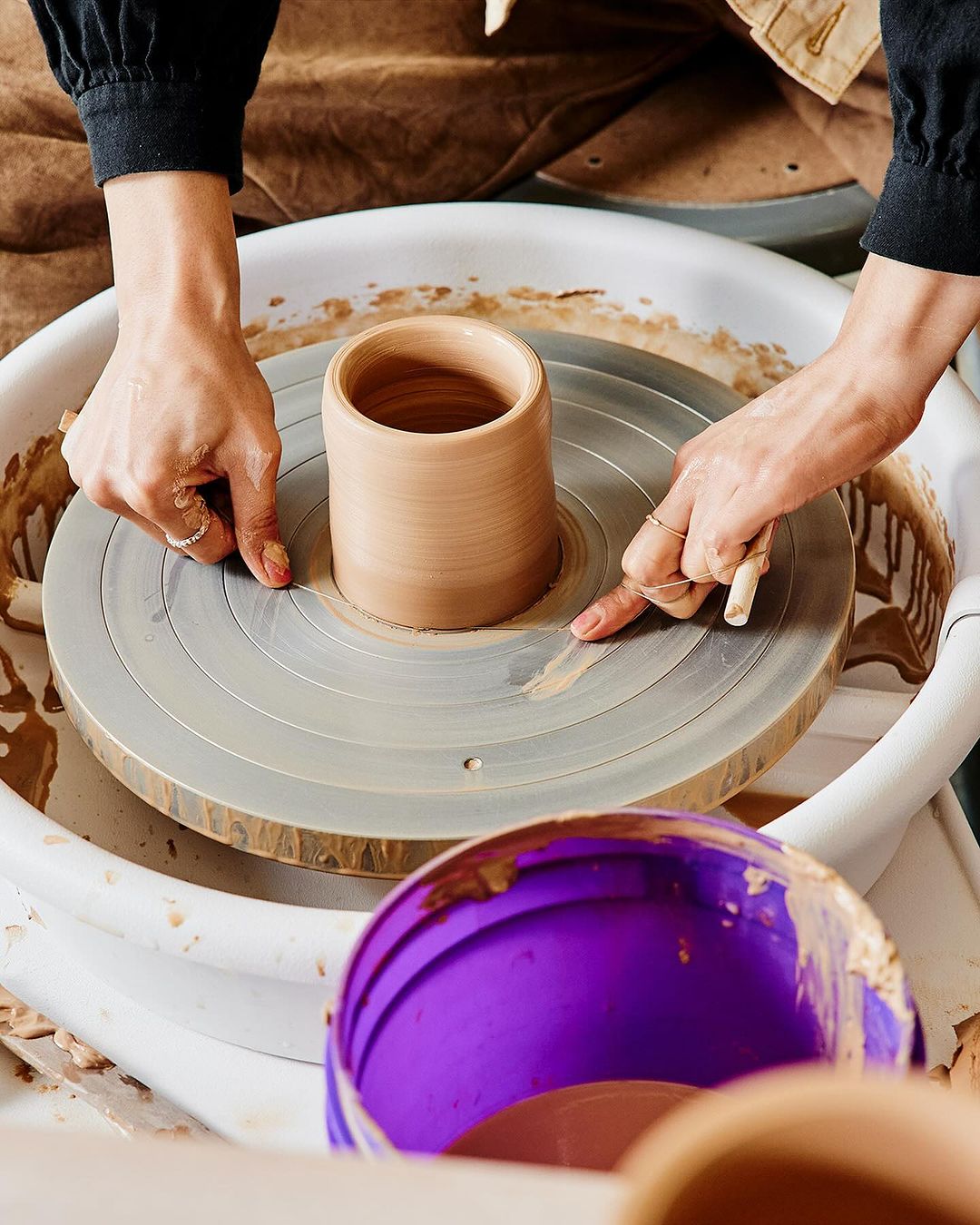 Adult female creating clay pot on pottery wheel in studio, shaping, and smoothing with her hands. Wheel, clay, and aprons visible.