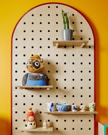 Eye-level shot of a pegboard shelf displaying a variety of ceramic art objects, pottery, a potted cactus, and yellow wall paint.