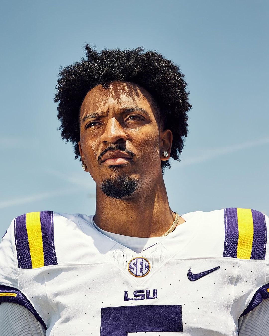 Portrait of a young adult football player with curly hair, wearing a white jersey with purple and gold details, posing against a blue sky.