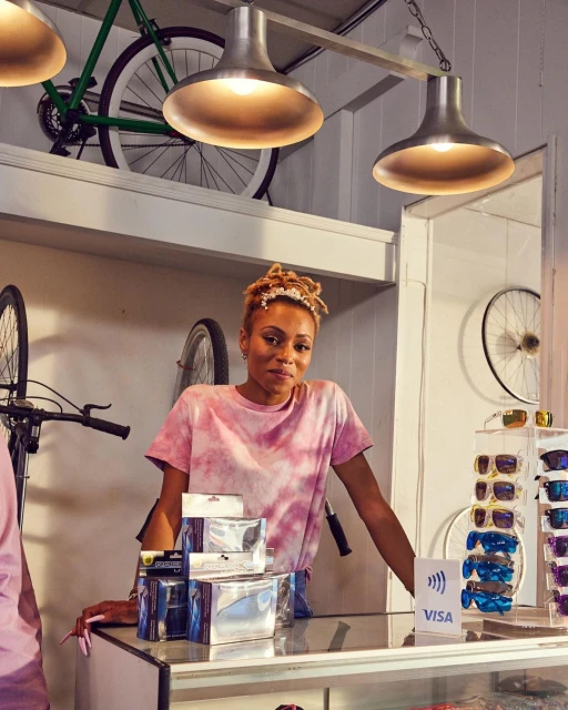 An adult female stands behind a counter in a bicycle shop, wearing a tie-dye t-shirt, with bicycles and accessories displayed.