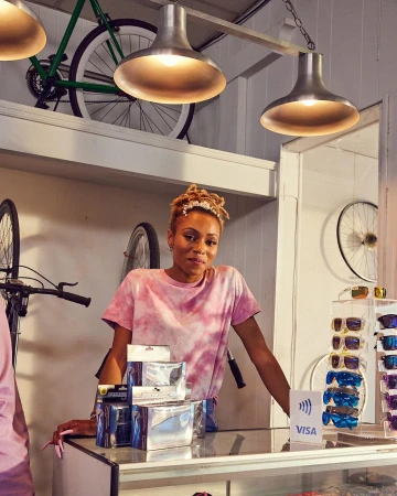 An adult female stands behind a counter in a bicycle shop, wearing a tie-dye t-shirt, with bicycles and accessories displayed.