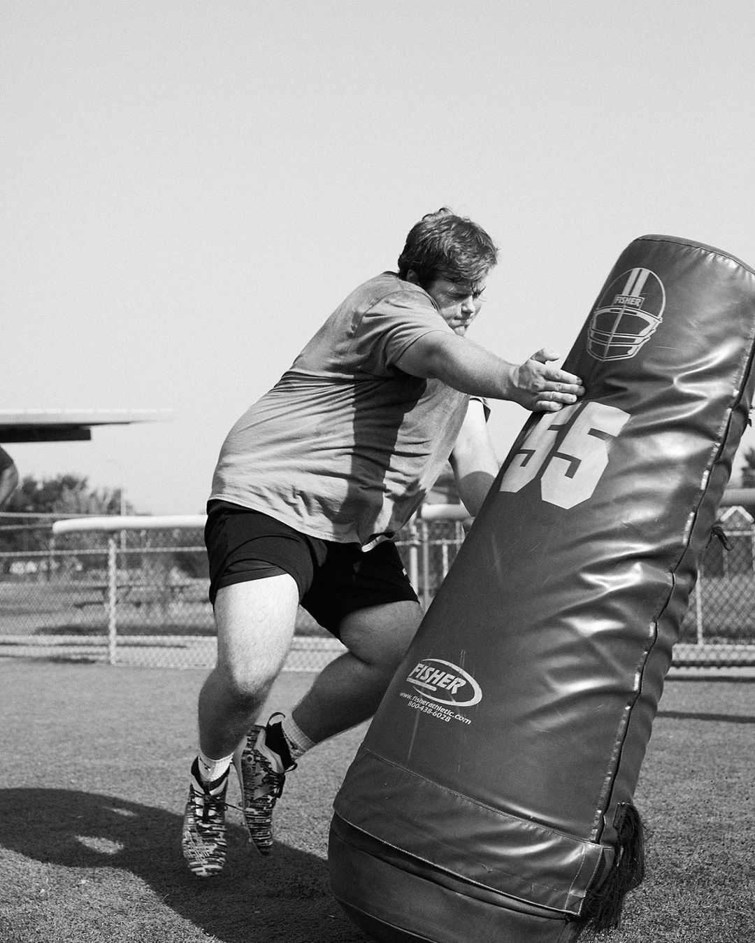 An adult male in sports clothing and shoes hits a football blocking pad on a field in black and white on a sunny day.