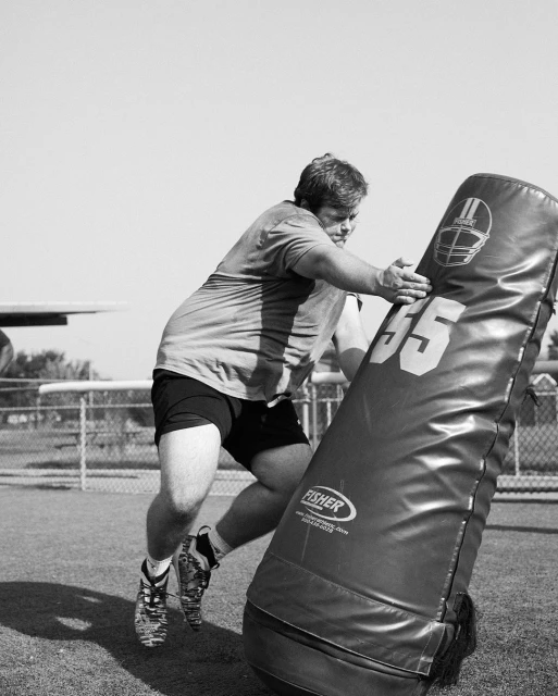 An adult male in sports clothing and shoes hits a football blocking pad on a field in black and white on a sunny day.