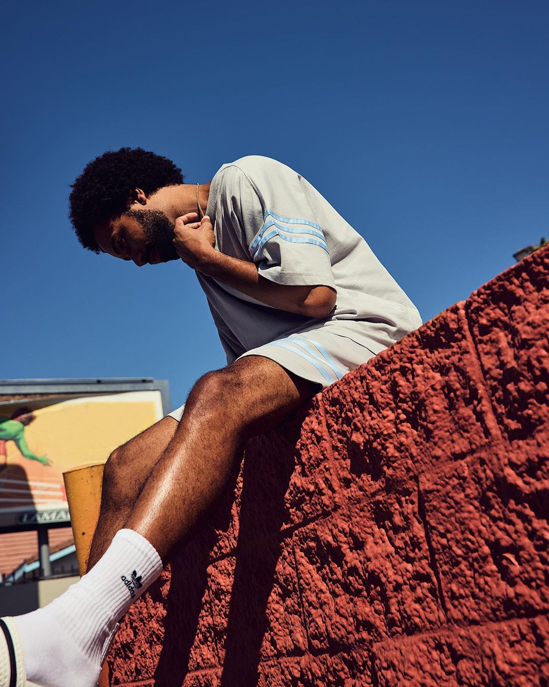 Adult male in gray athletic wear sits on an orange brick ledge under a blue sky, showcasing an outdoor urban fashion shot.