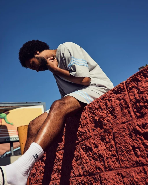 Adult male in gray athletic wear sits on an orange brick ledge under a blue sky, showcasing an outdoor urban fashion shot.