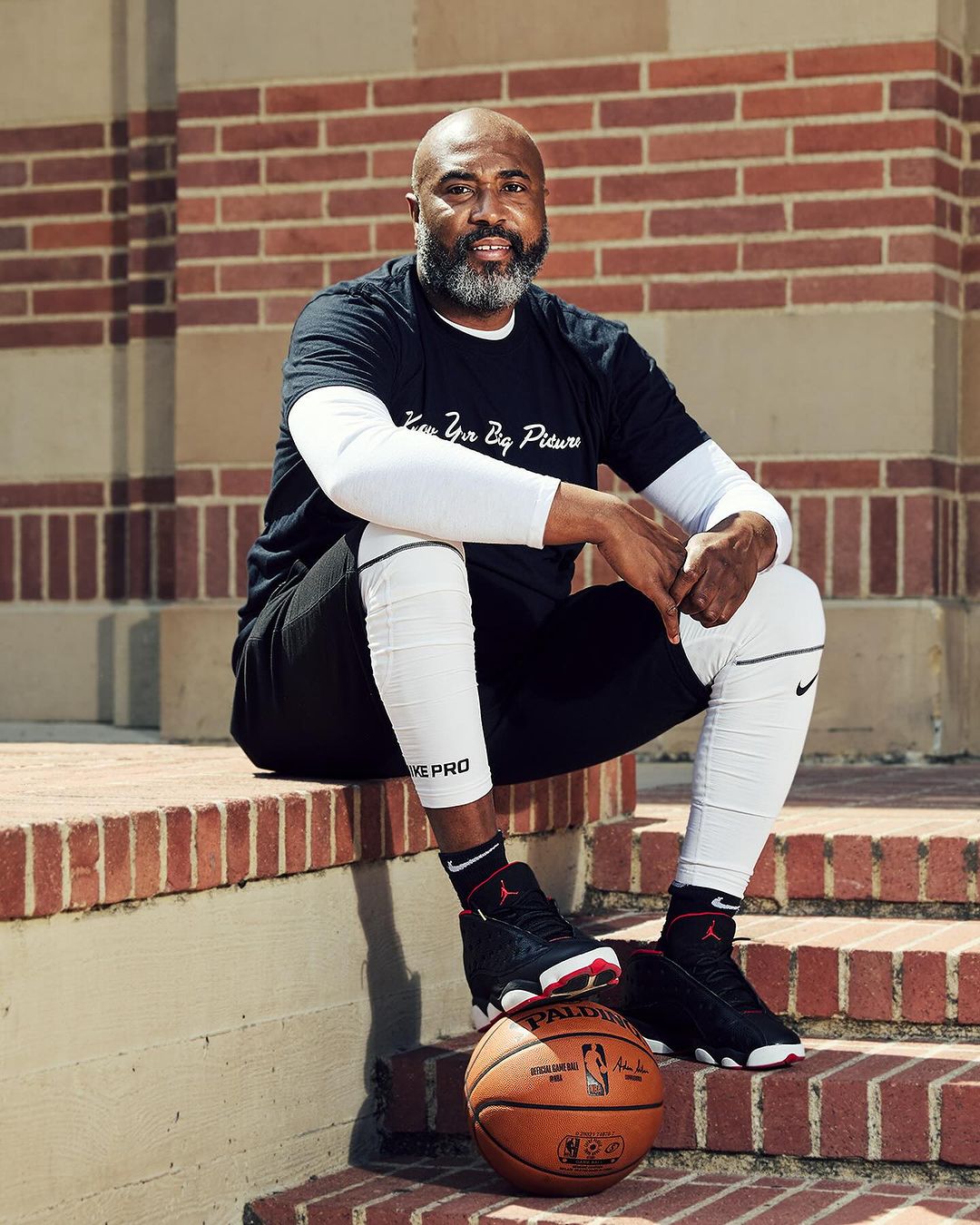 An African-American man sits on brick steps with a basketball. He wears a black shirt, white leggings, and black/red sneakers.