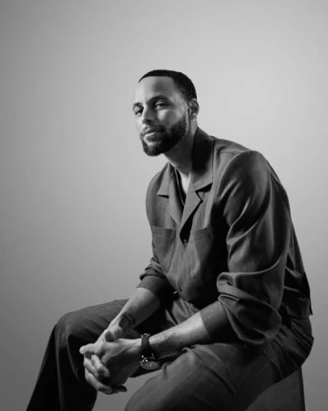 Black and white portrait of professional basketball player Stephen Curry seated in a collared shirt and trousers.