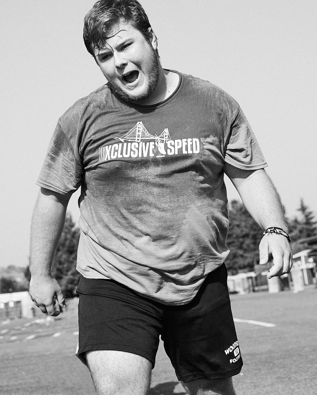 Black and white portrait of adult male in athletic wear outdoors with a graphic tee, standing on grass. Trees and sky in background.