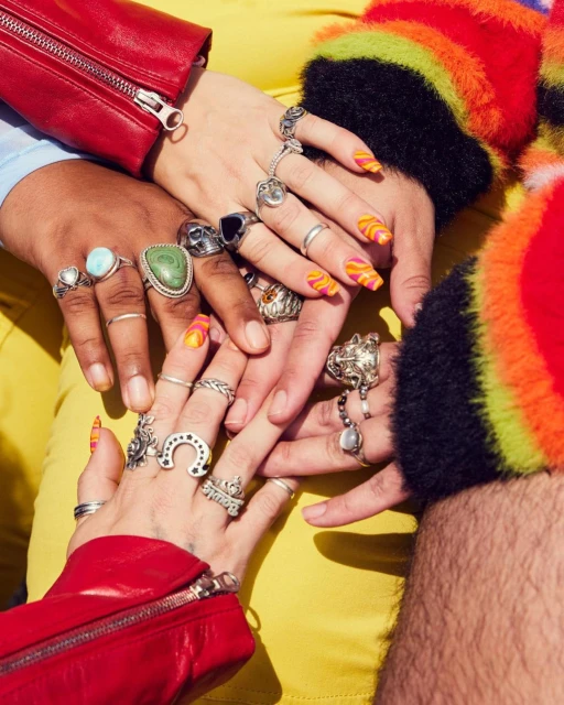 Close-up of three people's hands adorned with an assortment of silver rings and colorful nail art, set against a vibrant yellow backdrop.