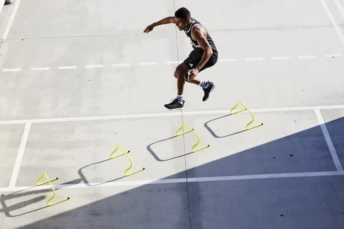 Athletic man in black sportswear leaps over yellow hurdles on a grey concrete surface, capturing a dynamic moment of fitness and agility.