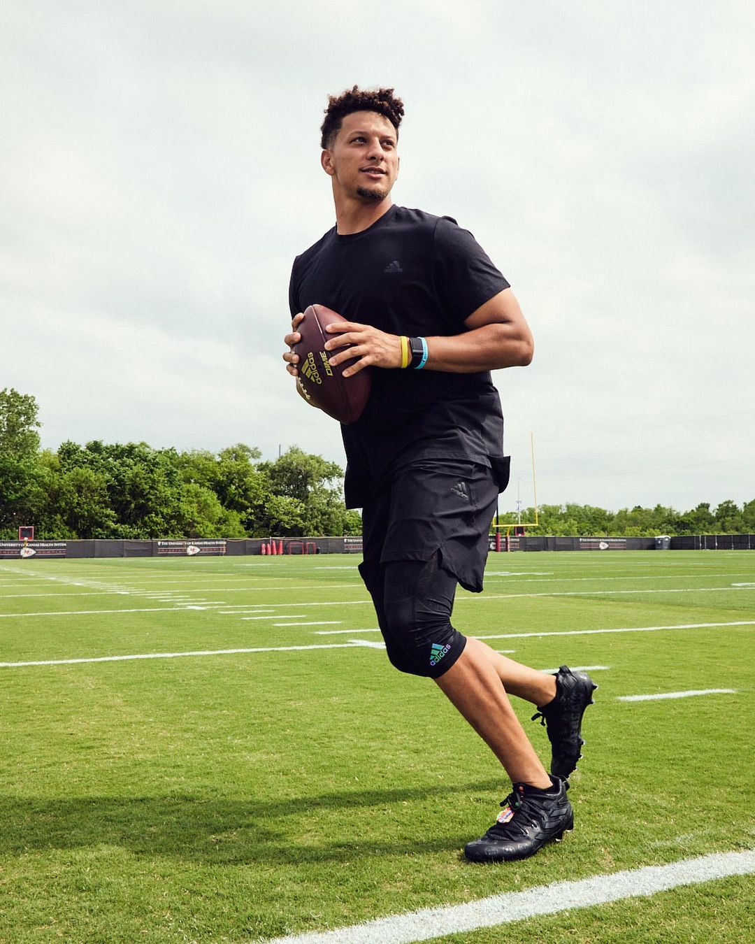 Male athlete in black athletic wear runs on a green sports field holding an American football. Clouds are visible in the sky.