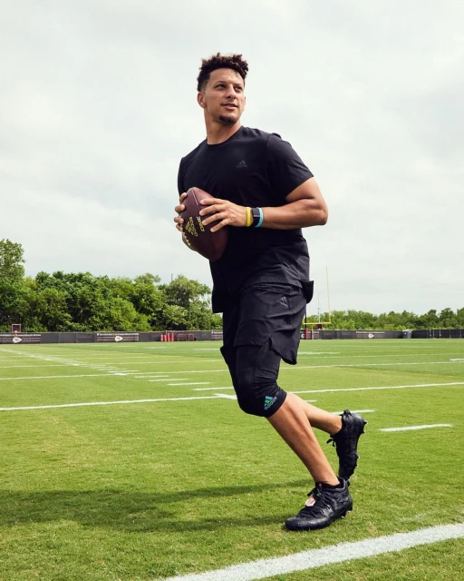 Male athlete in black athletic wear runs on a green sports field holding an American football. Clouds are visible in the sky.