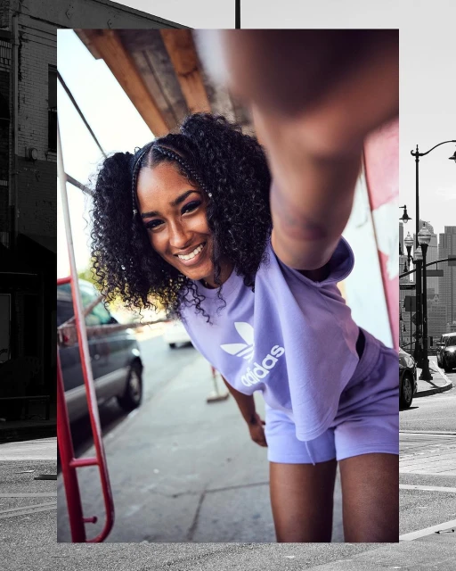 Smiling African-American woman takes selfie in lilac Adidas crop top and shorts against backdrop of city buildings and sidewalks.