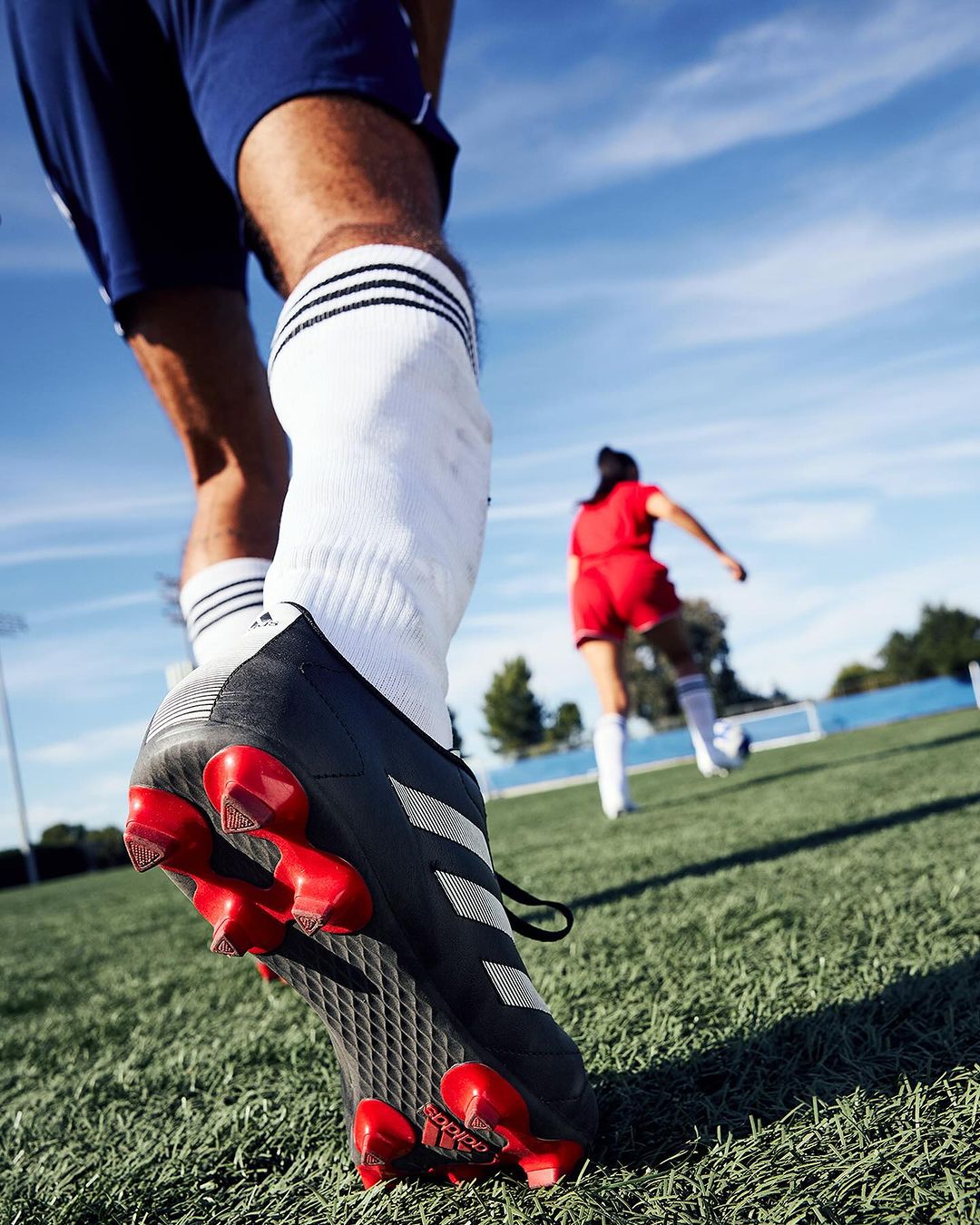 Low-angle shot of a soccer player's foot in a cleat with red studs, on a green field with a teammate in the background.