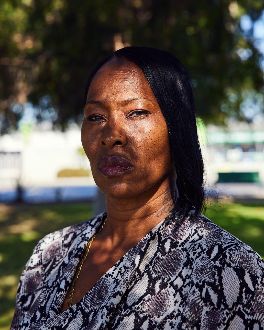 Close-up portrait of an adult woman with dark hair and a snakeskin print top in an outdoor setting with blurred green trees.