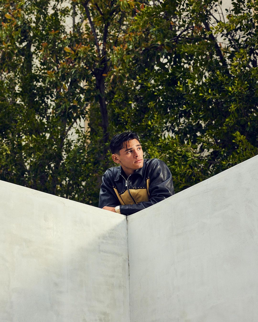 A young man wearing a black leather jacket leaning on a white concrete wall outdoors with trees behind