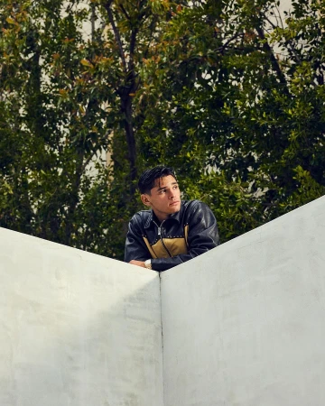 A young man wearing a black leather jacket leaning on a white concrete wall outdoors with trees behind