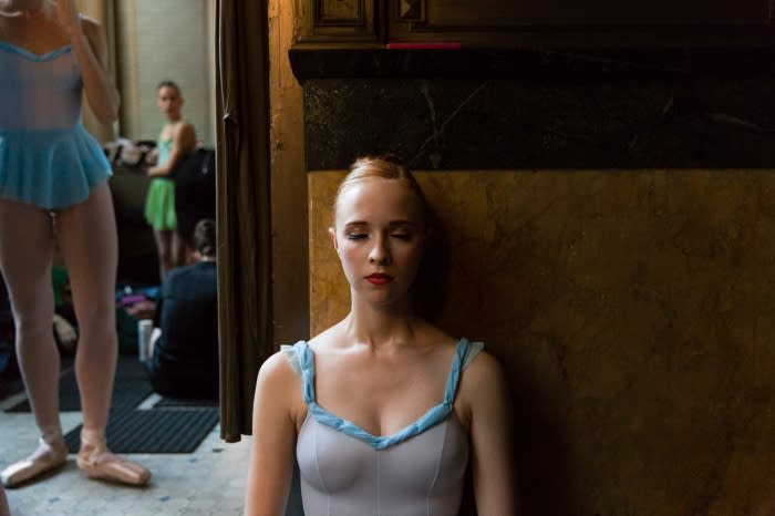 Portrait of a young woman in a white leotard and blue straps and skirt, with other dancers in the background and a dark wood wall.