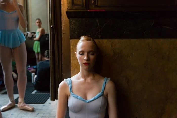 Portrait of a young woman in a white leotard and blue straps and skirt, with other dancers in the background and a dark wood wall.