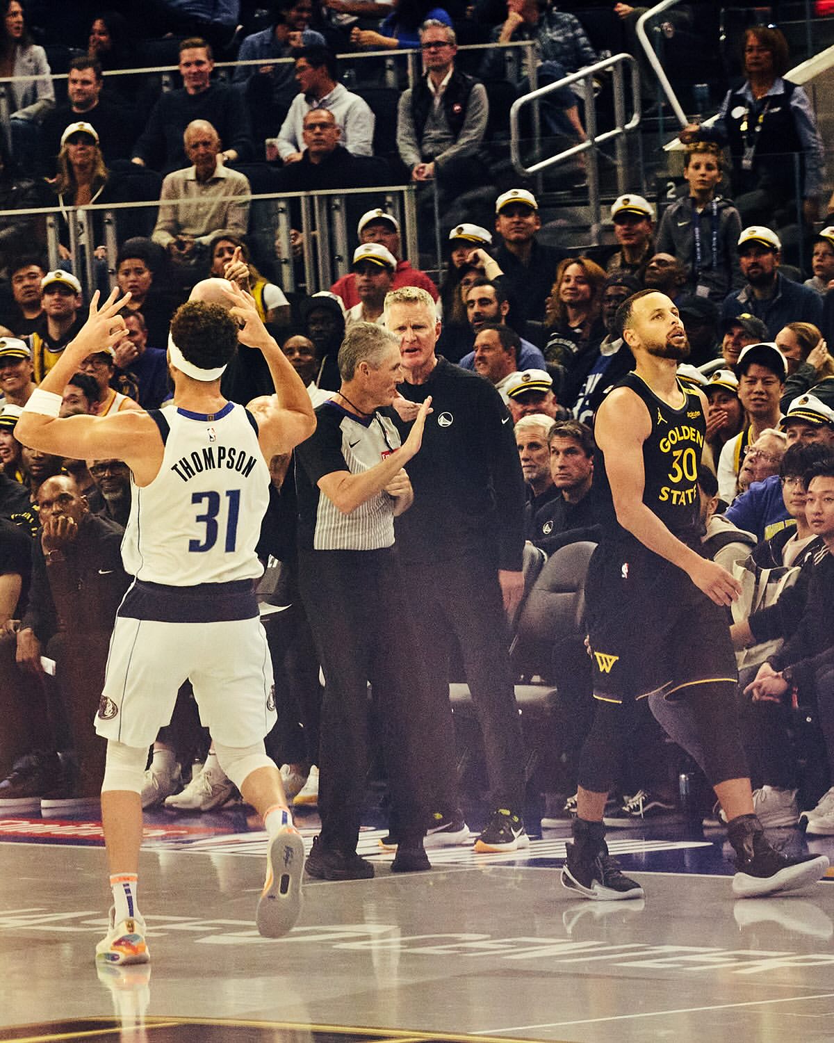 Action shot on a basketball court: player in white raising arms, referee in center, Curry in Warriors jersey, crowd background.