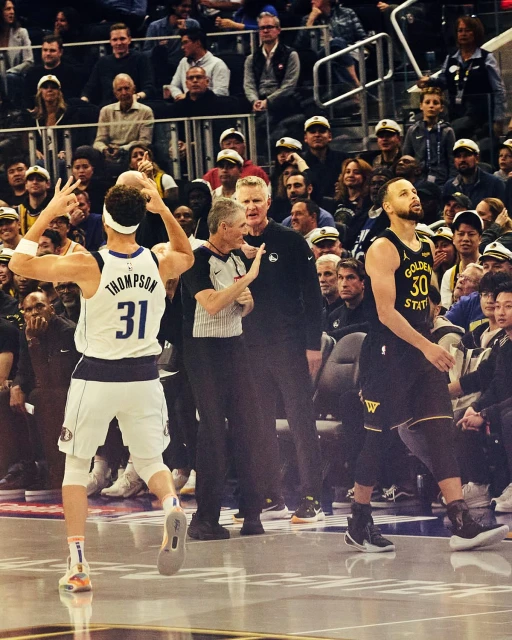 Action shot on a basketball court: player in white raising arms, referee in center, Curry in Warriors jersey, crowd background.