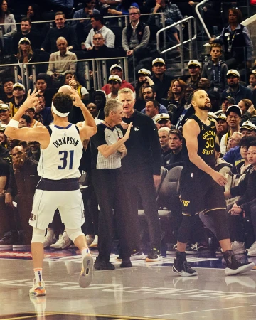 Action shot on a basketball court: player in white raising arms, referee in center, Curry in Warriors jersey, crowd background.