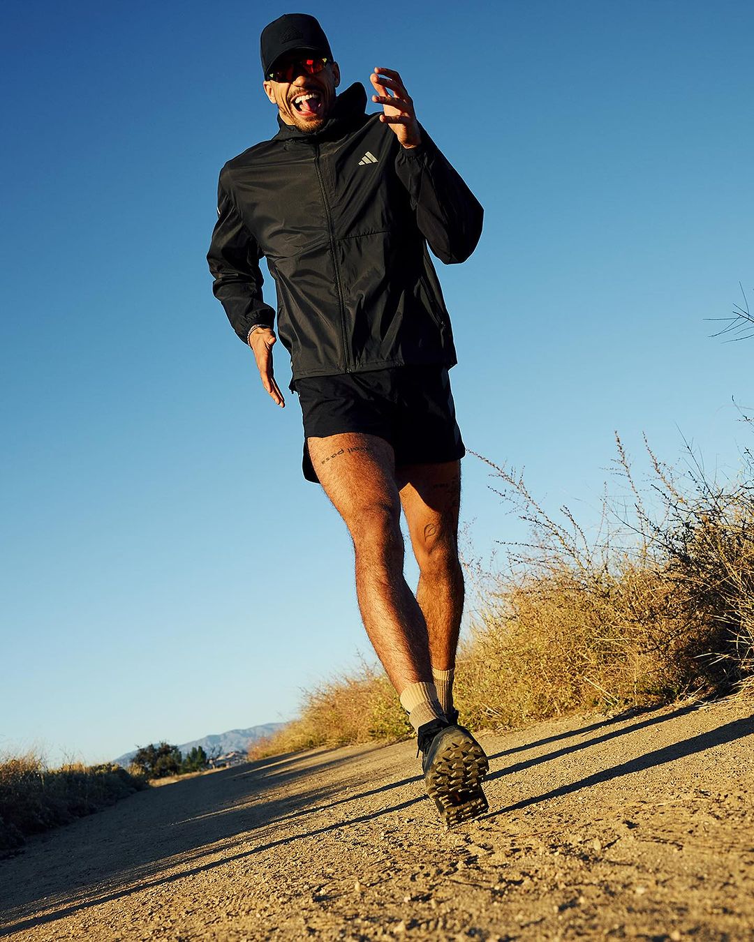 An adult man is running on a dirt trail wearing a black jacket, shorts, sunglasses, and a baseball cap against a blue sky.