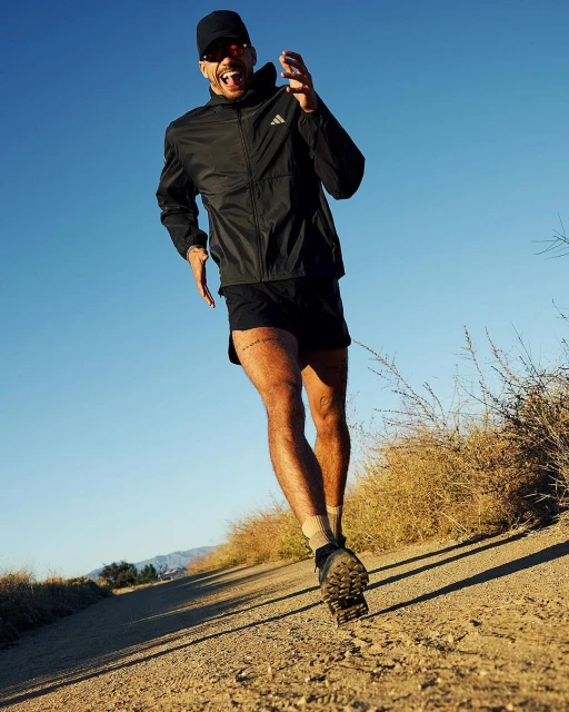 An adult man is running on a dirt trail wearing a black jacket, shorts, sunglasses, and a baseball cap against a blue sky.
