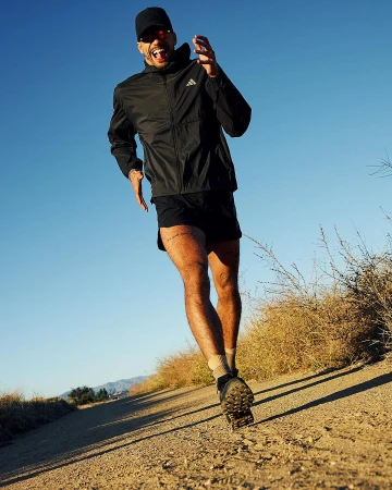 An adult man is running on a dirt trail wearing a black jacket, shorts, sunglasses, and a baseball cap against a blue sky.