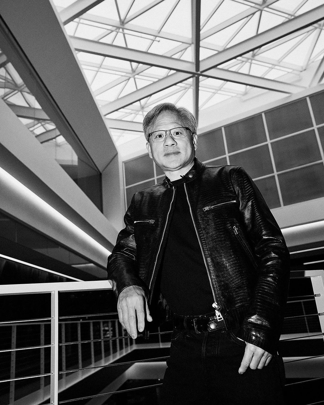 Black and white portrait of a mature adult man in glasses and a leather jacket standing indoors against an architectural backdrop.