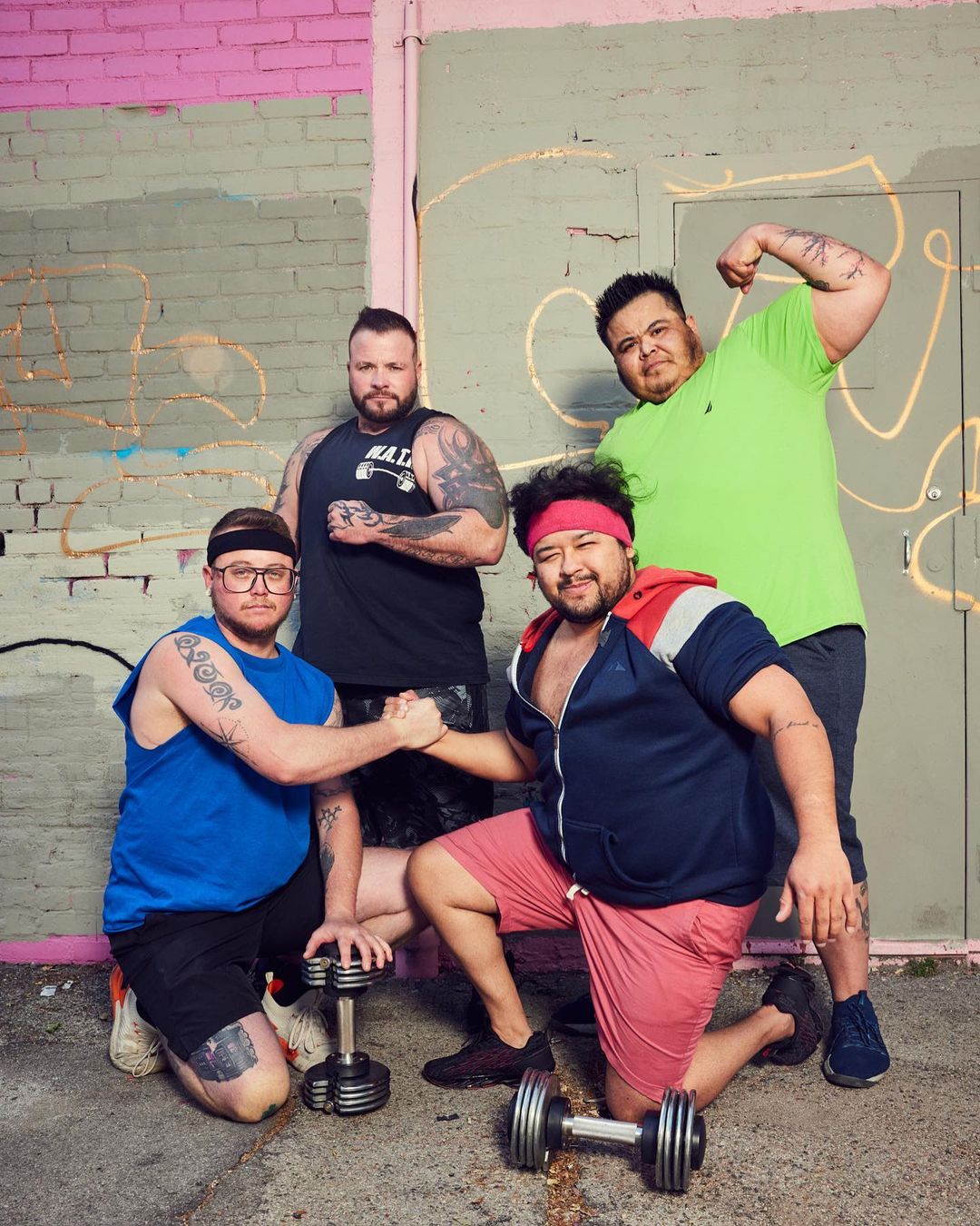 Four athletic men pose outdoors in workout attire with dumbbells. Two men are kneeling with dumbbells, two men are standing.