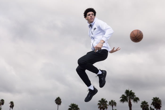 A man in athletic wear jumps in the air to throw a basketball, with palm trees visible in the background under a cloudy sky.