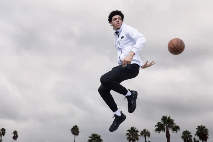 A man in athletic wear jumps in the air to throw a basketball, with palm trees visible in the background under a cloudy sky.