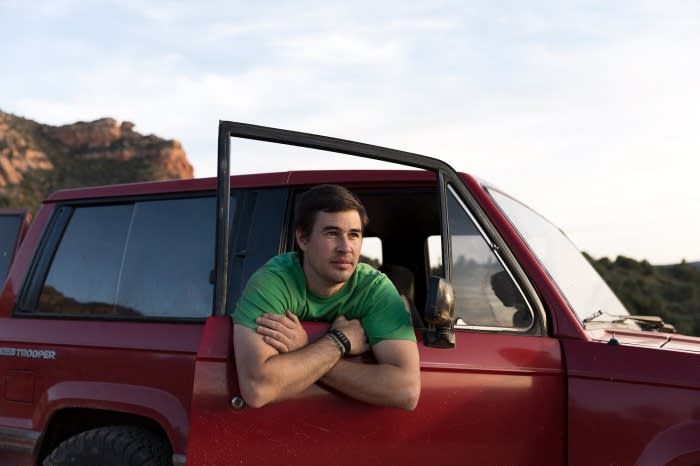 Man in a red SUV looking out the open door, arms crossed. Outdoor shot with clear sky and distant rock formation.
