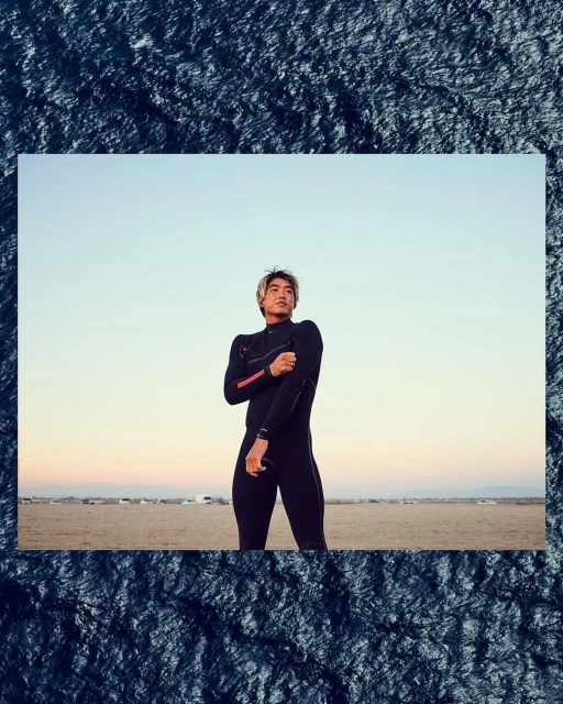 Asian male in wetsuit standing on a sandy beach under a blue sky. He is warming up his arms, and there is natural lighting.