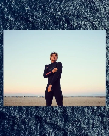 Asian male in wetsuit standing on a sandy beach under a blue sky. He is warming up his arms, and there is natural lighting.