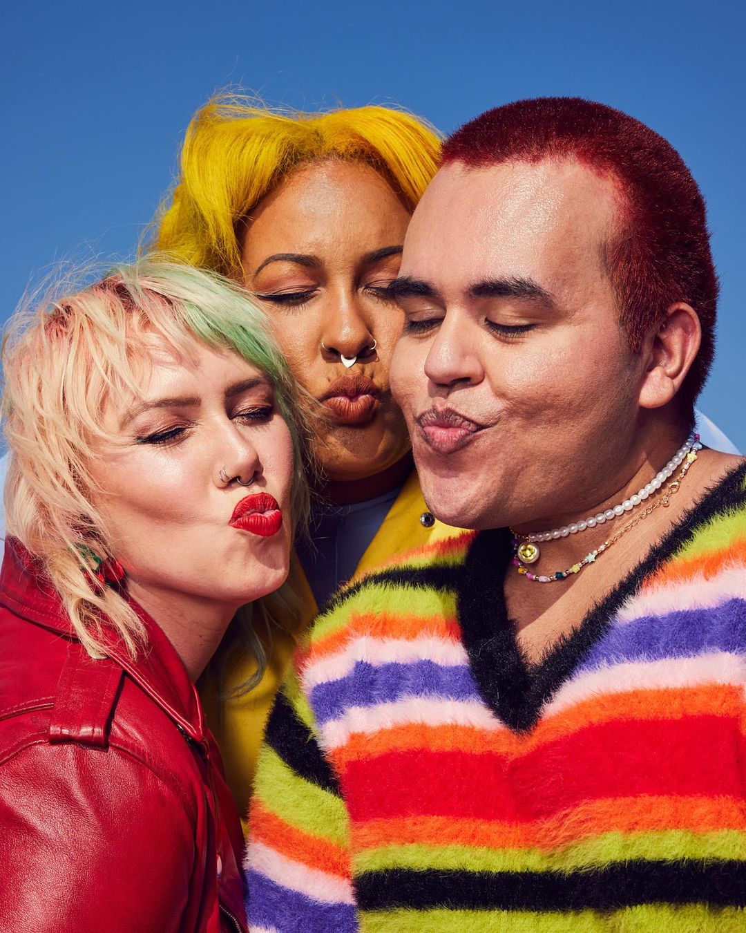Three people with colorful hair and stylish outfits are posing closely together with pouting lips against a clear, blue sky backdrop.