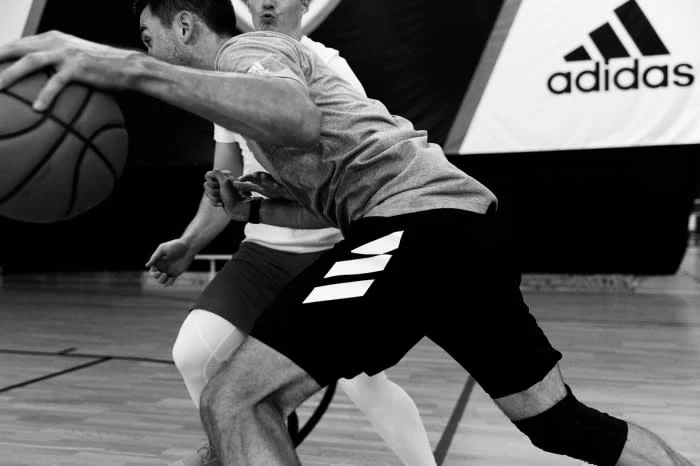 Two athletes in action with a basketball on a basketball court, sporting athletic wear with the Adidas logo in the background.