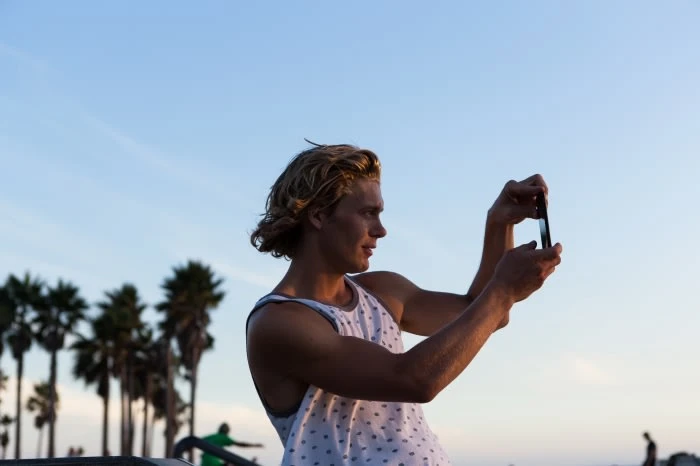 A young man takes a photo with his cell phone outside in the sunshine, with palm trees and a blue sky in the background.