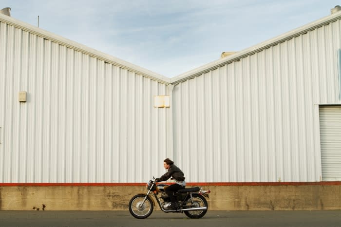 Man riding a motorcycle past a large white industrial building exterior