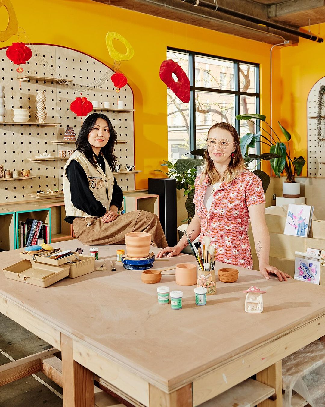 Two women sit and stand around a large craft table with pottery, paints, and brushes inside an indoor craft studio with yellow walls.