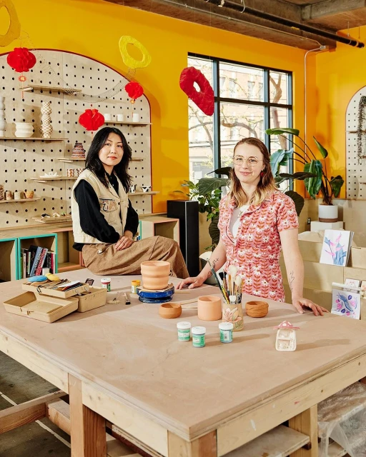 Two women sit and stand around a large craft table with pottery, paints, and brushes inside an indoor craft studio with yellow walls.