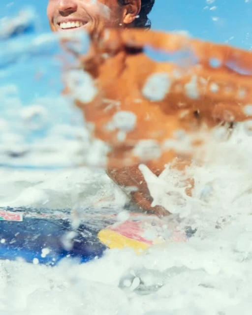Adult male surfing on blue board, smiling with sunlight and sea spray creating a dynamic, energetic image.
