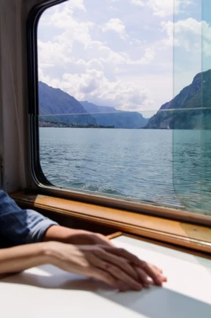 Scenic lake view seen from a train window with a woman's hands resting on a white table near the window, creating a travel-inspired image.