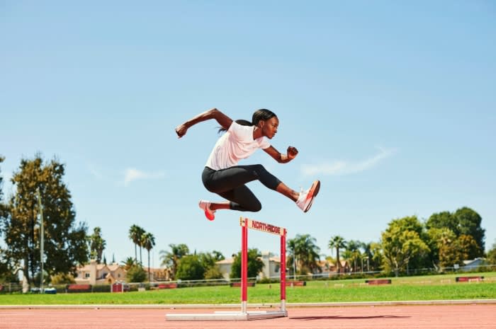 An athletic woman jumps over a hurdle on a track on a sunny day wearing running clothes and shoes, with trees and blue sky background.