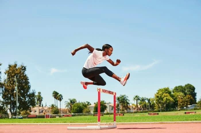 An athletic woman jumps over a hurdle on a track on a sunny day wearing running clothes and shoes, with trees and blue sky background.