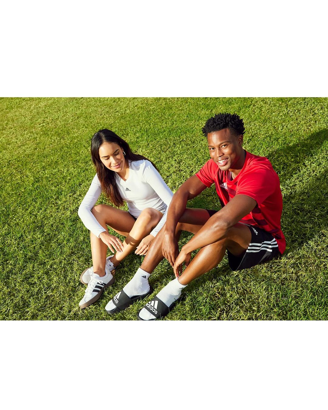 Two people sit on grass wearing Adidas apparel. An adult man and woman relax outdoors, facing the camera, with green grass around them.