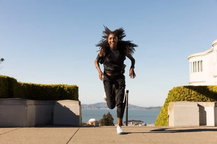 Athletic African-American woman running outdoors on a sunny day. She is wearing sportswear with ocean and city skyline in background.